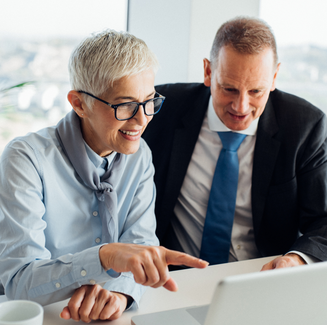 older white woman smiling looking at computer with glasses on next to white man with suit and tie on